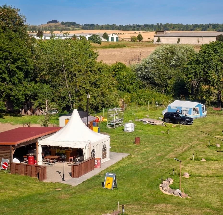 Weißes Pavillonzelt und überdachter Sitzplatz mit Tischen am Rand der Campingwiese, rechts ein Zelt mit Auto, im Hintergrund Felder